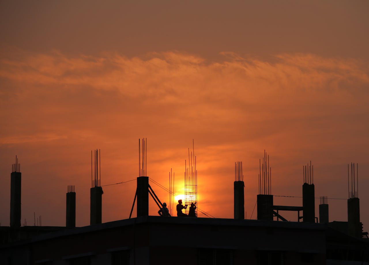 hero-02 Silhouette of workers on a construction site against a vibrant sunset sky.